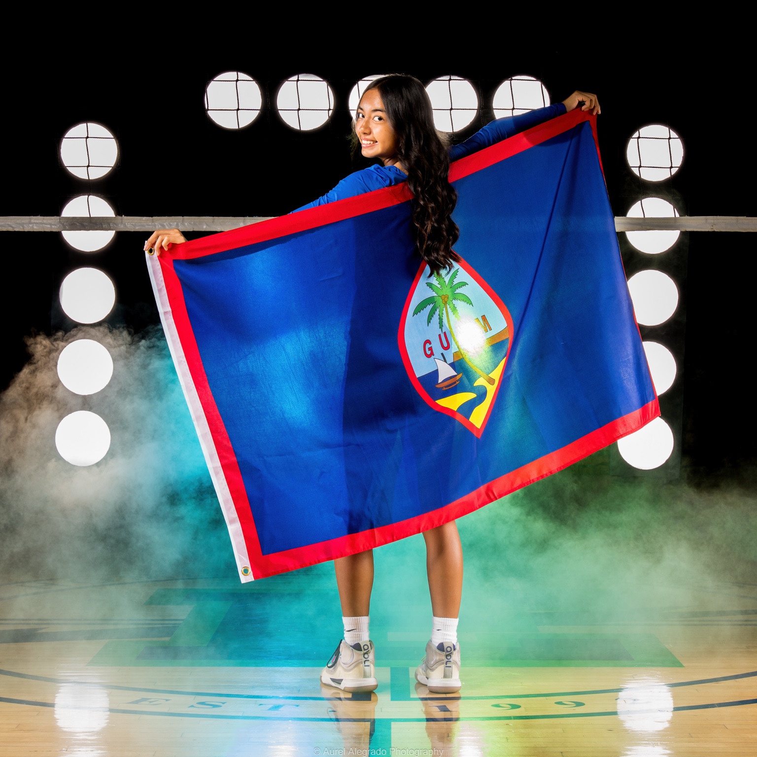 Kaylia looking back over her shoulder while holding the flag of Guam open behind her on a lit gym floor