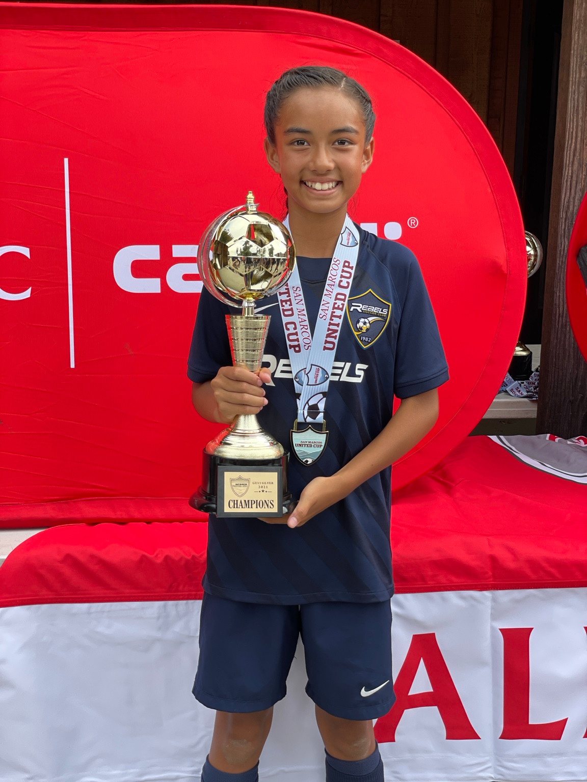Younger Kaylia in a navy soccer kit holding a gold trophy and wearing a medal