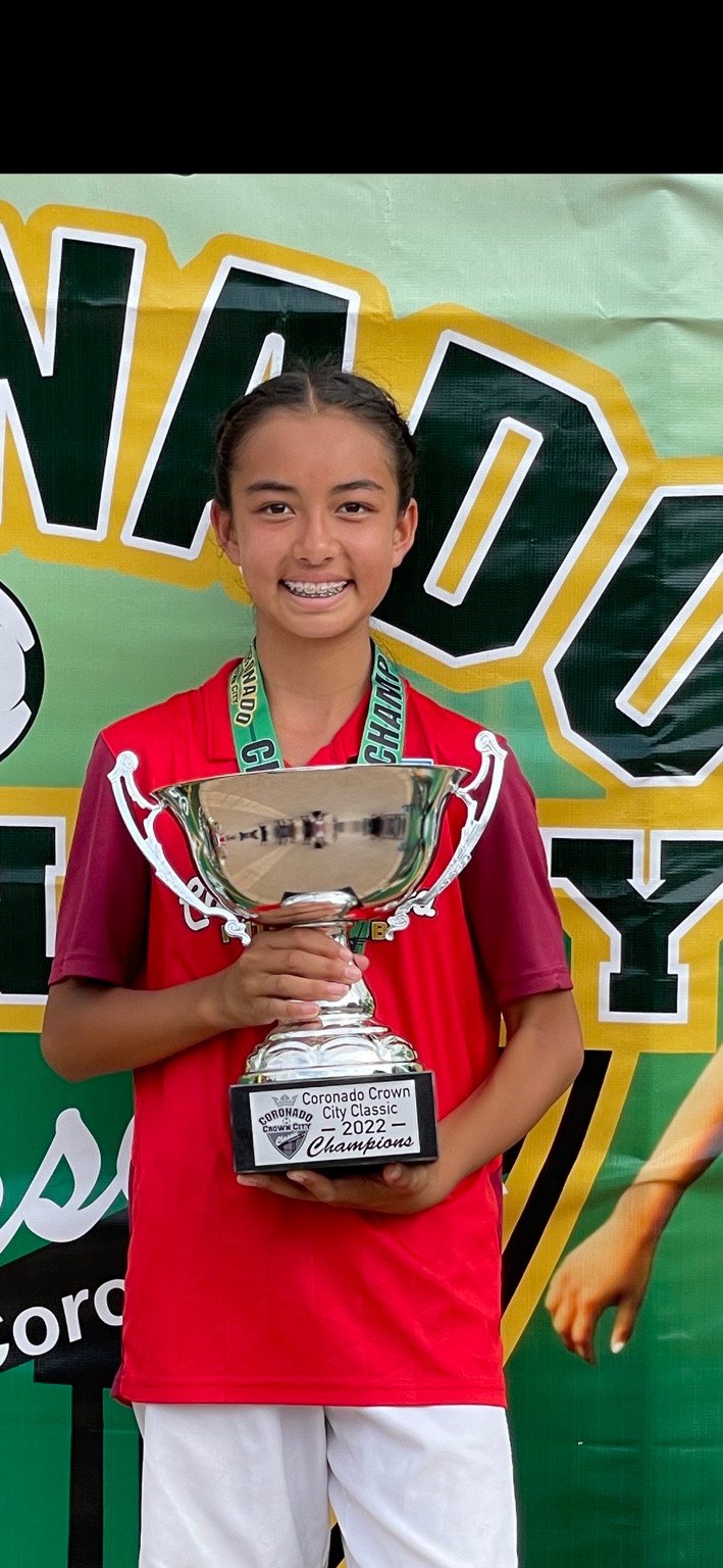 Younger Kaylia in a red soccer jersey holding a silver cup trophy