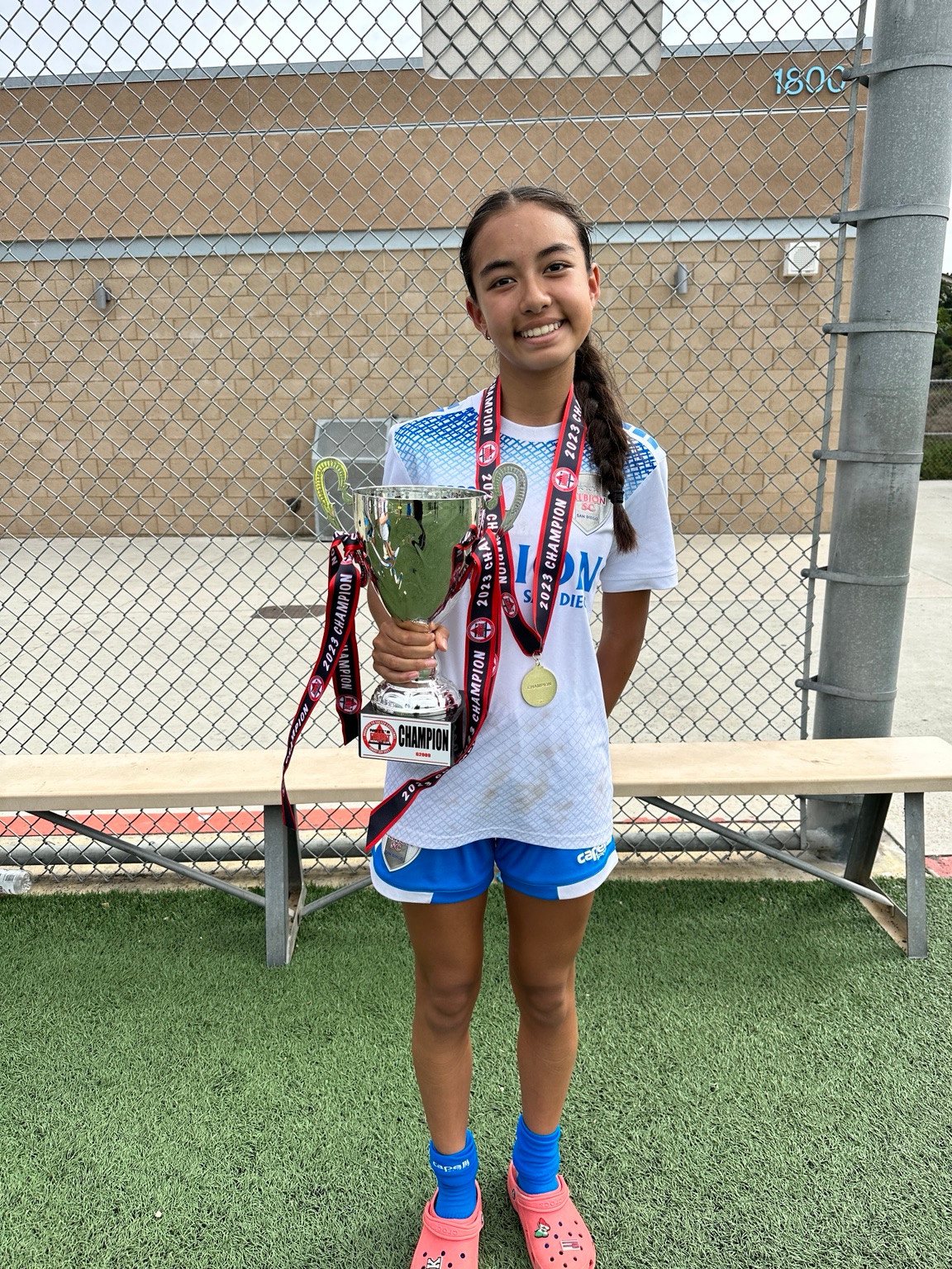 Kaylia in a white soccer kit holding a silver cup with a medal around her neck beside a field